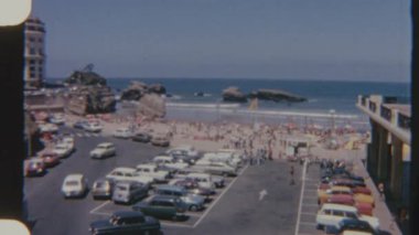 Biarritz sahil beldesinde içi araba dolu bir otopark kumlu bir sahile bakıyor. İnsanlar güneşli bir günün tadını çıkarıyor ve 8 mm 'lik bir filme çekiliyor..