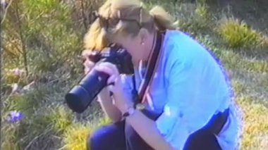 Nostaljik bir VHS klibi, bahçeye çömelmiş, parlak çiçekler ve yeşillikler arasında bir telefoto lensi ile yabani çiçeklerin yakın plan fotoğraflarını çeken sıradan giyinmiş bir kadını gösteriyor.