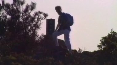 Sırt çantası ve kamerası olan bir kadın tepede siluetlenir, doğa tarafından çerçevelenir ve günbatımında parlayan pastel gökyüzü