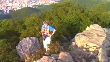 Kalitesiz kaliteli bir video, sırtında sırt çantası ve kamerasıyla kayalık bir dağ zirvesini keşfeden, sahil şehrine ve açık mavi gökyüzünün altındaki limana bakan bir kadını gösteriyor.