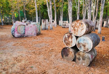 Heyecanlı oyuncuların saklandığı paintball sahasında eski paslı fıçılar ve hasarlı lastikler.