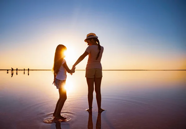 Två vackra systrar går längs spegelsjön på kvällen eldig solnedgång — Stockfoto