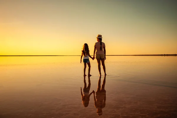 Duas irmãs encantadoras caminham ao longo do lago espelho no pôr-do-sol ardente da noite — Fotografia de Stock