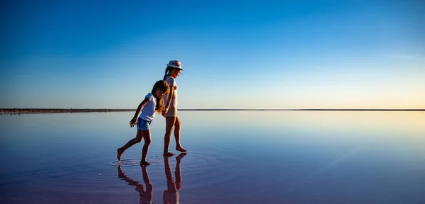 İki küçük mutlu kız kardeş ayna gölünde yürüyorlar sıcak akşam güneşinin tadını çıkarıyorlar. — Stok fotoğraf