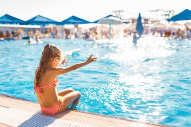 Sevimli küçük bir kız havuzun kenarında oturuyor ayakları suyun içinde güneşin tadını çıkarıyor.