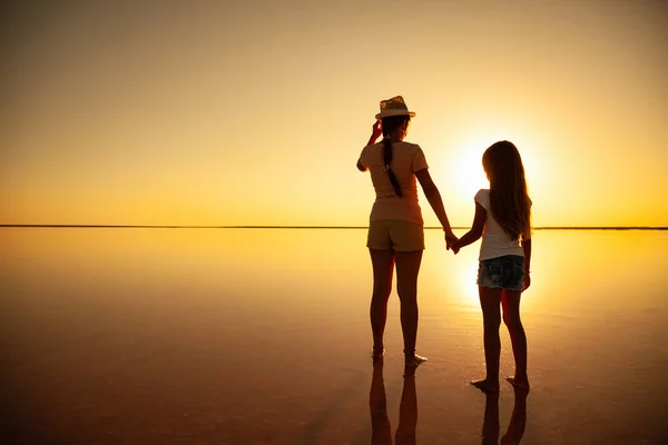 Two lovely sisters walk along the mirror lake in the evening fiery sunset — Stock Photo, Image