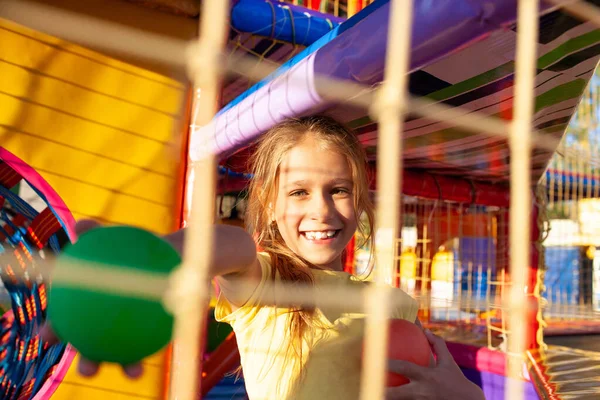 Мила смішна дівчина сидить на ігровому майданчику з м'яким і яскравим обладнанням і кидає барвисті кульки в бік камери — стокове фото