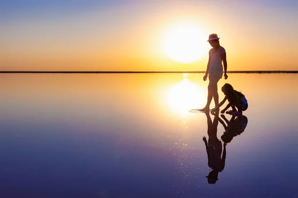 Дві милі сестри ходять уздовж дзеркального озера ввечері вогненного заходу сонця — стокове фото