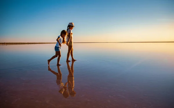 İki küçük mutlu kız kardeş ayna gölünde yürüyorlar sıcak akşam güneşinin tadını çıkarıyorlar. — Stok fotoğraf
