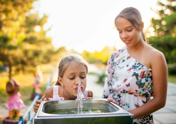 Zwei lustige fröhliche, wunderbare Schwestern trinken frisches Wasser aus einem kleinen Brunnen in einem sommerlich warmen, sonnigen Park — Stockfoto