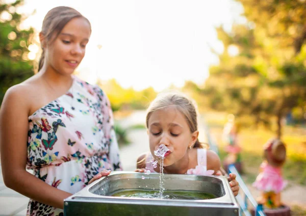 Due divertenti allegre meravigliose sorelle bevono acqua fresca da una piccola fontana in un parco estivo caldo e soleggiato — Foto Stock