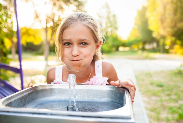 Allegro ragazza meravigliosa beve acqua fresca da una piccola fontana in un parco estivo caldo e soleggiato — Foto Stock