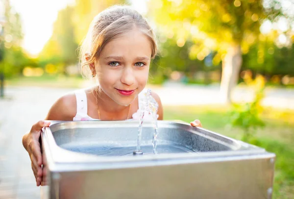 Allegro ragazza meravigliosa beve acqua fresca da una piccola fontana in un parco estivo caldo e soleggiato — Foto Stock