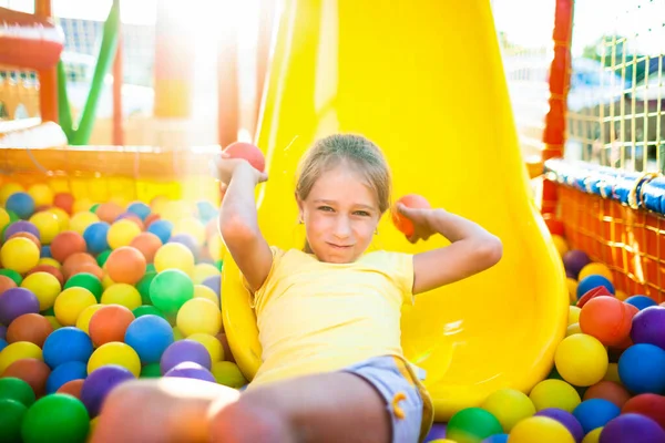 Мила весела дівчина лежить на дитячому майданчику з м'яким і яскравим обладнанням і кидає різнокольорові кульки в камеру — стокове фото