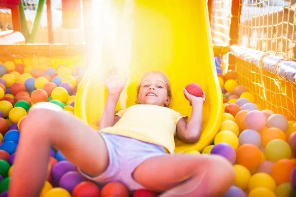 Мила весела дівчина лежить на дитячому майданчику з м'яким і яскравим обладнанням і кидає різнокольорові кульки в камеру — стокове фото