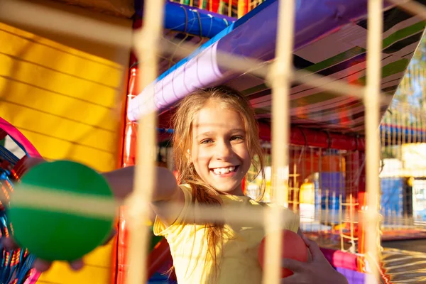 Мила смішна дівчина сидить на ігровому майданчику з м'яким і яскравим обладнанням і кидає барвисті кульки в бік камери — стокове фото