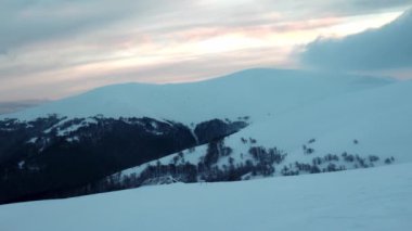 Kardan bir halı ve köknar ormanlarıyla kaplı büyük tepeler bulutlu bir gökyüzü ve günbatımının arka planına karşı.