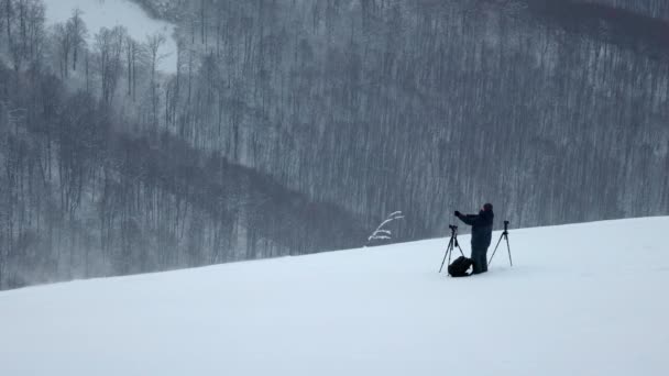 Un fotógrafo con dos cámaras en trípodes en una tormenta de nieve se encuentra en una colina y tiene una vista de un bosque — Vídeo de stock