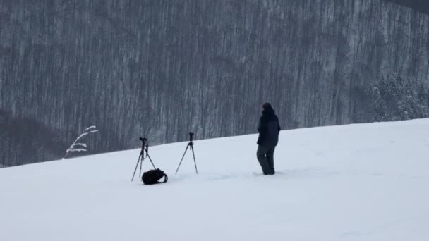 Фотограф с двумя камерами на штативах в снежную бурю стоит на холме и смотрит на лес — стоковое видео