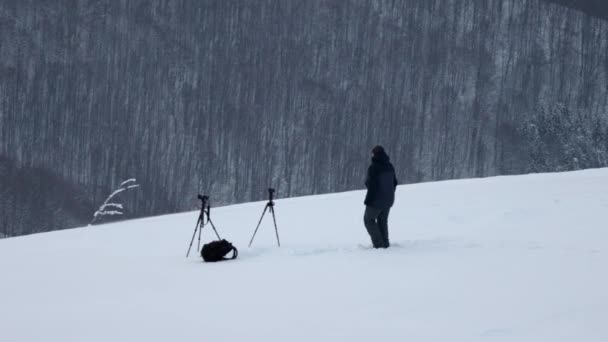 Een fotograaf met twee camera 's op statieven in een sneeuwstorm staat op een heuvel en kijkt uit op een bos — Stockvideo