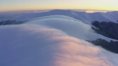 Bulut dalgaları yavaşça kar kaplı dağların tepelerinden akar. Soğuk bir günbatımında ladin ormanlarıyla kaplıdır.