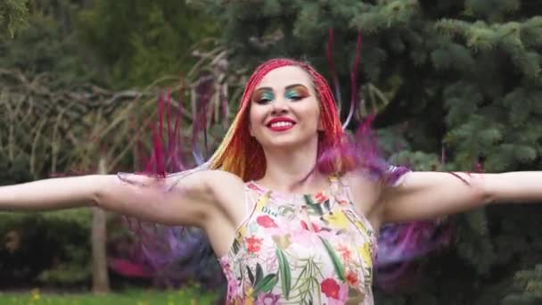 Una chica con una sonrisa feliz en un vestido con trenzas africanas y giros de maquillaje y arroja su pelo en el parque lluvioso de primavera — Vídeo de stock