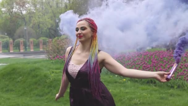 O fată cu un zâmbet cu machiaj strălucitor și împletituri colorate într-o rochie violet suflă fum violet artificial într-un parc — Videoclip de stoc