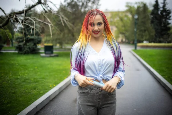 A girl with a confident look in a pale blue shirt with bright glitter makeup and artificial colored braids. Posing against the backdrop of a blooming park on spring day and confidently looking ahead.