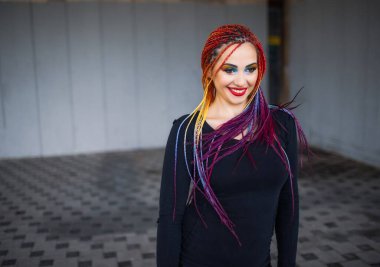 Parlak makyajlı ve çok renkli yapay örgülü siyah şık elbiseli en güzel kız. Güzel daireler çiziyor ve eski bir binanın arka planında uzun örgüler sallıyor..