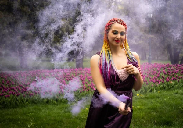 Smiling girl in purple satin dress with long multi-colored braids and eye-catching glitter makeup. Unusual artificial smoke of purple color covers the girl in the spring park.