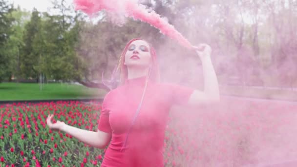 Uma menina com maquiagem com tranças de arco-íris em vestido vermelho posando em fumaça vermelha contra o fundo de um canteiro de flores florescendo — Vídeo de Stock