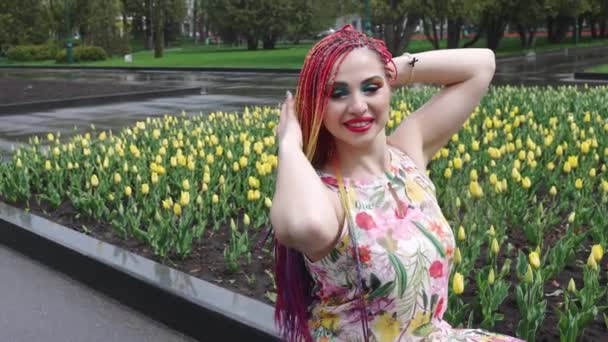 Una chica con maquillaje brillante y trenzas de arco iris africano está disfrutando de la llegada de la primavera en el parque floreciente en clima cálido — Vídeos de Stock