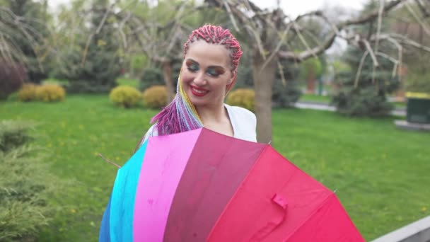 Una ragazza carina con trecce multicolori e trucco luminoso in una camicia bluastra in posa con un ombrello arcobaleno — Video Stock