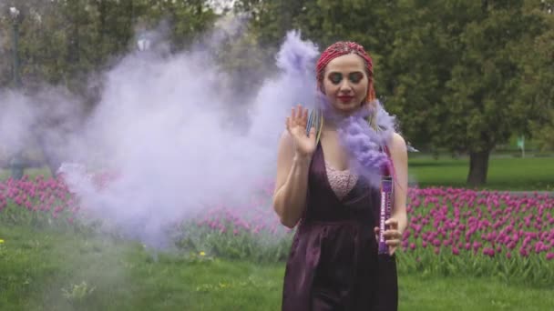 Una ragazza con un sorriso con trucco luminoso e trecce colorate in un abito viola soffia fumo viola artificiale in un parco — Video Stock