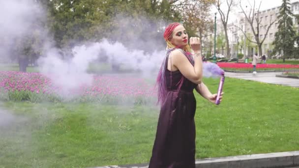 O fată cu un zâmbet cu machiaj strălucitor și împletituri colorate într-o rochie violet suflă fum violet artificial într-un parc — Videoclip de stoc