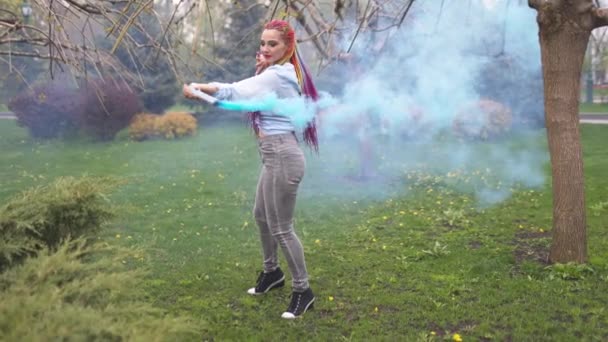 Hermosa chica con trenzas africanas multicolores posando con humo azul sobre el fondo del parque verde — Vídeos de Stock