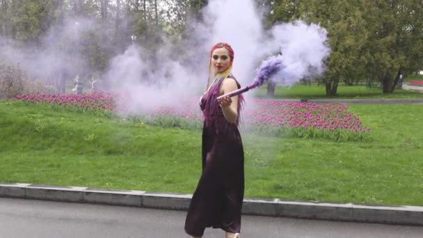 Una chica con una sonrisa con maquillaje brillante y trenzas de colores en un vestido púrpura sopla humo púrpura artificial en un parque — Vídeos de Stock