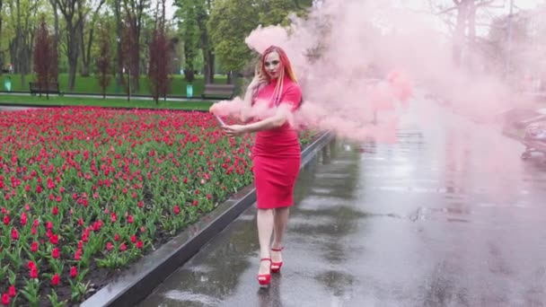 Uma menina com maquiagem com tranças de arco-íris em vestido vermelho posando em fumaça vermelha contra o fundo de um canteiro de flores florescendo — Vídeo de Stock