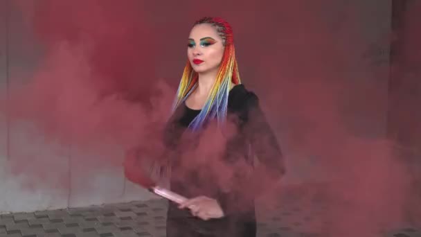 暖かい春の街で赤い濃い煙の中でポーズをとる、色の編組と虹のメイクを持つシンプルな黒のドレスの女の子 — ストック動画