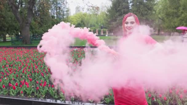 Uma menina com maquiagem com tranças de arco-íris em vestido vermelho posando em fumaça vermelha contra o fundo de um canteiro de flores florescendo — Vídeo de Stock