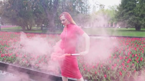 A lány smink szivárvány zsinór piros ruhában pózol a vörös füst a háttérben egy virágzó virágágyás — Stock videók