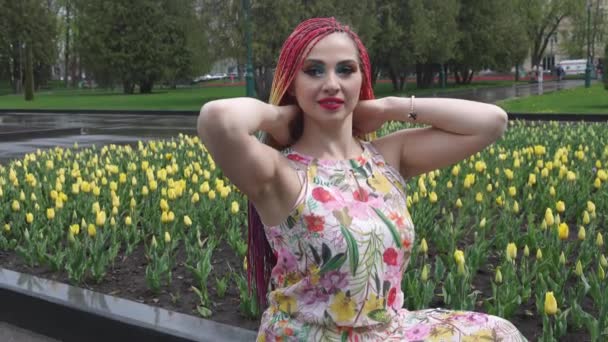 Uma menina com maquiagem brilhante e tranças de arco-íris Africano está desfrutando a chegada da primavera no parque florescente em tempo quente — Vídeo de Stock