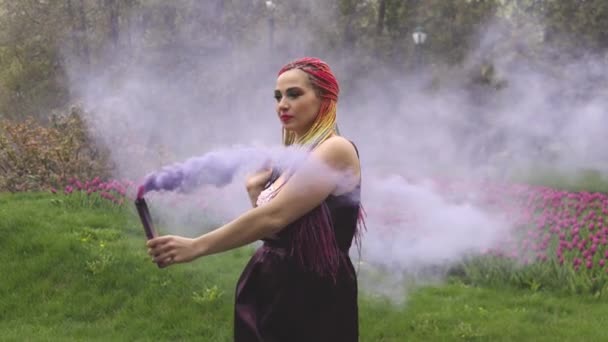 Una chica con una sonrisa con maquillaje brillante y trenzas de colores en un vestido púrpura sopla humo púrpura artificial en un parque — Vídeos de Stock