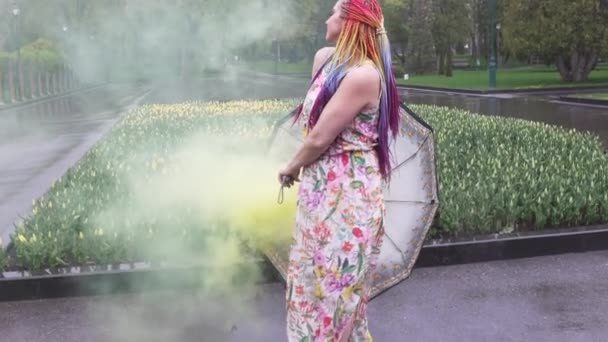 Chica con trenzas africanas y maquillaje en vestido de primavera está bailando bajo la lluvia con paraguas en humo amarillo en un parque de la ciudad — Vídeos de Stock