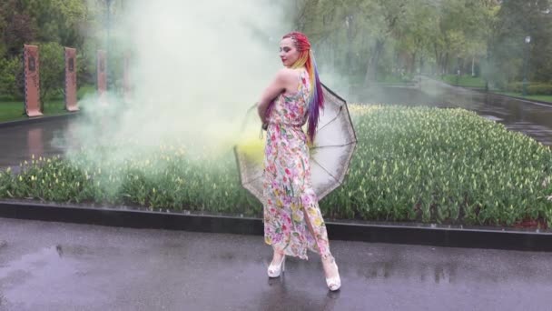 Menina com tranças africanas e maquiagem em vestido de primavera está dançando na chuva com guarda-chuva em fumaça amarela em um parque da cidade — Vídeo de Stock