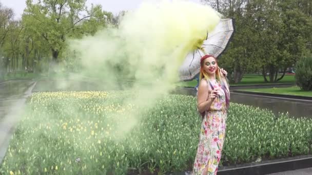 Ragazza con trecce africane e trucco in abito primaverile sta ballando sotto la pioggia con ombrello in fumo giallo in un parco cittadino — Video Stock