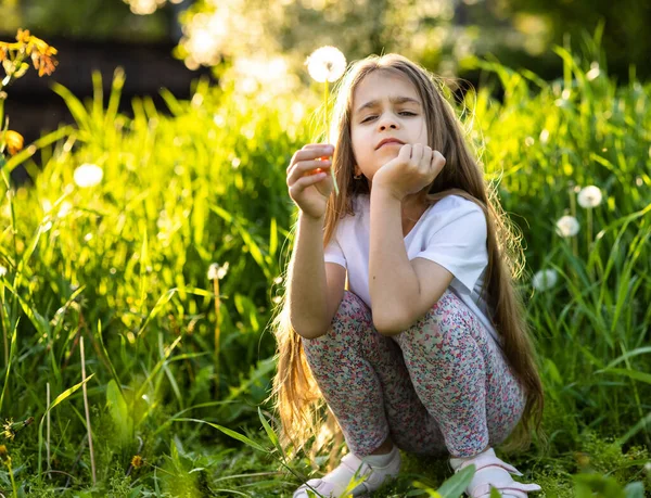 Ein Mädchen blickt konzentriert in die Kamera und hält einen weißen Löwenzahn auf einem Hintergrund aus grünem Gras im Garten — Stockfoto