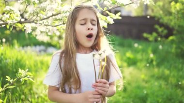 Bir kız kameraya dikkatle bakar ve bahçedeki yeşil çimlerin üzerinde beyaz bir karahindiba tutar.