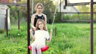 Hafif giysili neşeli çocuklar yeşil bir bahçede salıncakta sallanıyor ve mutlu bir gülümsemeyle gülüyorlar.