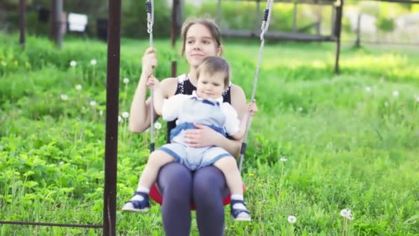 Sød bror og ældre søster ride på en lys swing på varm forårsdag på baggrund af en grøn blomstrende have – Stock-video